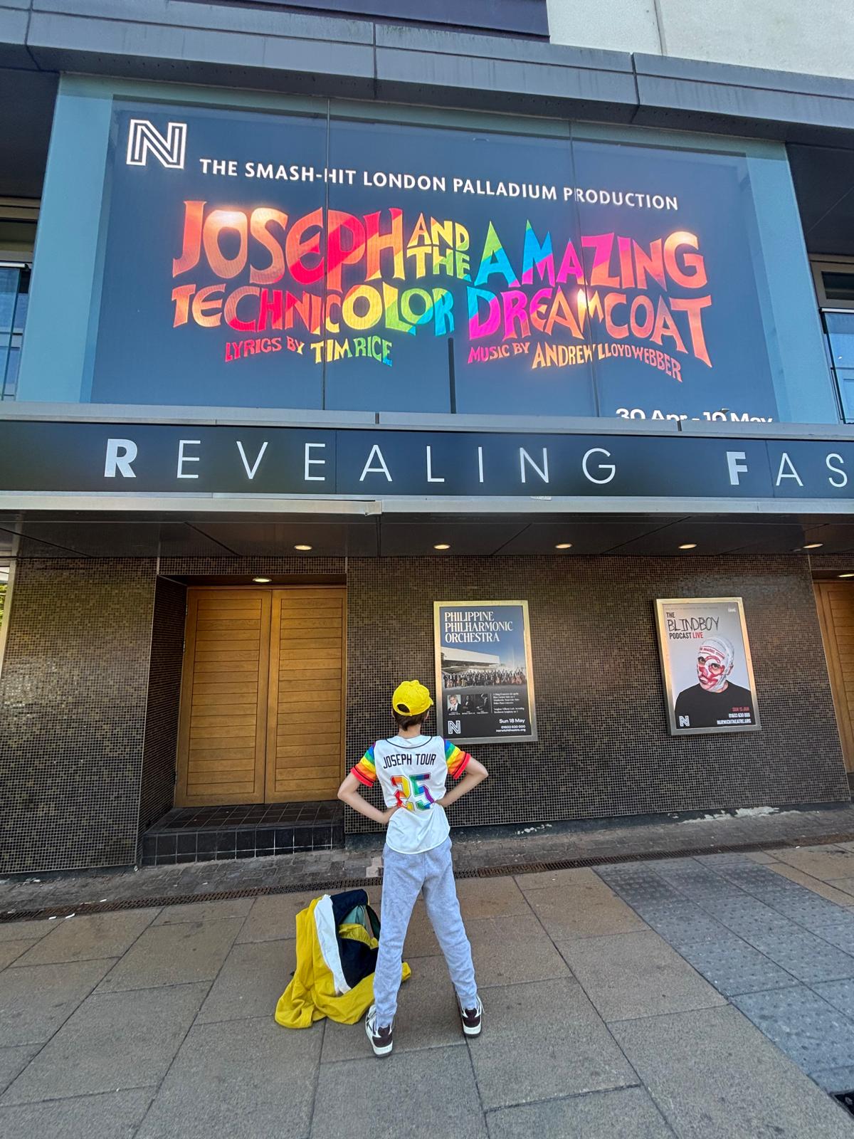 A young performer seen from behind, wearing a Joseph tour shirt, looking up at the theatre billboard for Joseph and the Amazing Technicolor Dreamcoat