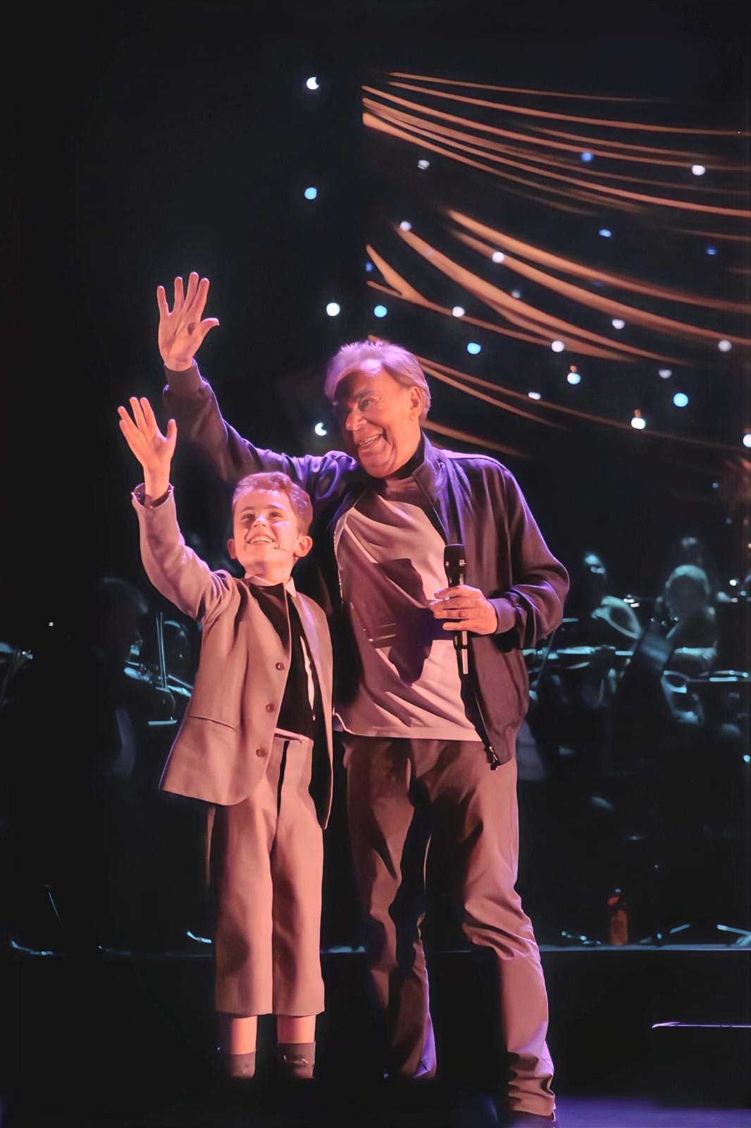 A young KTTA performer waving to the audience on stage alongside Andrew Lloyd Webber